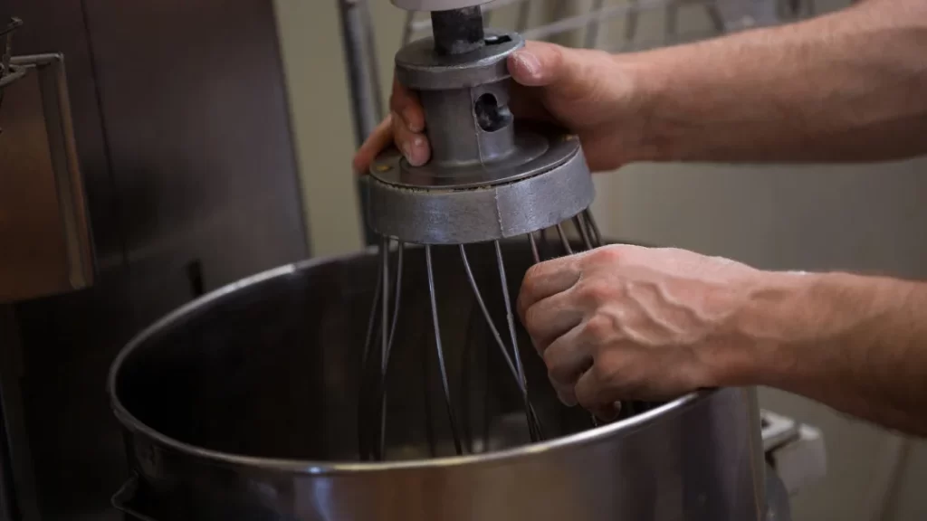 A person mixing ingredients in a large metal bowl, using a bakery mixer for preparation.